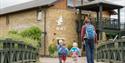 A man and two children, all with backpacks, walk toward the London Wetland Centre building. The atmosphere is inviting and family-friendly.