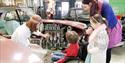A guide showing a family a car at British Motor Museum. the family features a small child in a manual wheelchair
