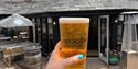 A hand with teal nail polish holds a pint of beer labeled "Wooda Cornwall" outside a rustic pub with stone architecture and patio seating. Bright, sun