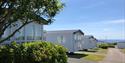 Row of white mobile homes with large windows, set among green bushes and trees on a sunny day. Ocean view under a clear, blue sky in the background.