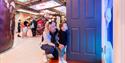 a toddler is looking through a blue door with his mother kneeled behind him.