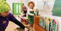 a member of staff is kneeling down scrubbing a wicker basket whilst a young girl plays with an interactive wooden display of flowers.