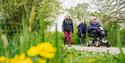 Three adults, one using a mobility scooter, walk on a path beside vibrant greenery and yellow flowers, conveying a peaceful, inclusive outdoor setting