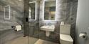 Modern luxury bathroom with gray marble walls, glass walk-in shower, floating sink, illuminated mirrors, and white toilet in a serene setting.
