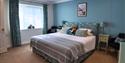 Cosy blue bedroom with a neatly made bed, striped gray throw, floral pillows, bedside tables, framed art, and softly filtered window light.