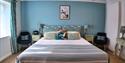 Bright, inviting bedroom with blue walls, a neatly made striped bed, patterned pillows, matching lamps, dark armchairs, and framed art.