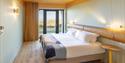 Inviting hotel room with a cosy bed, colorful blanket, and soft lighting. Large window reveals a serene outdoor view, creating a warm, tranquil ambian