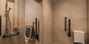Accessible restroom with beige tiles, featuring a shower area with grab bars and foldable seat, alongside a toilet with support rails.