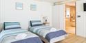 Two neatly made twin beds with blue and white striped bedding in a bright room. Adjacent bathroom, light wood floors, two framed art pieces above. Cos