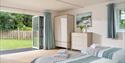 Bright bedroom with two neatly made beds and folded towels, open patio doors revealing a lush garden. Light blue curtains and calming decor create a s