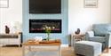 A cozy living room featuring a blue accent wall with a wall-mounted TV above an electric fireplace. A wooden coffee table displays flowers, cups, and