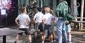 A group of children in uniforms and a staff member stands outdoors, observing a playful statue. The scene conveys curiosity and engagement on a sunny