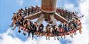 People enjoying a thrill ride, suspended in mid-air on a circular swing against a bright blue sky. Their expressions show excitement and thrill.