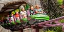 People on a roller coaster ride with green and purple cars. They are laughing and excited as the ride emerges from a wooden tunnel. Lush greenery surr