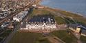 Aerial view of a coastal town featuring a grand white building with a clock tower near the sea, surrounded by green fields and a residential area. The