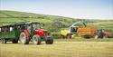 A red tractor pulls a green trailer with people through a field, alongside a harvester and another tractor. Lush hills and clear skies are in the back