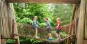 Three children play joyfully on a red rope bridge between wooden structures in a forest. They balance and smile, surrounded by lush green trees.