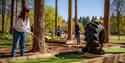 A woman plays mini-golf in a forest-themed course with large trees and a tractor tire obstacle. People enjoy a sunny day outdoors in the background.