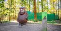 Statue of the Gruffalo, a brown fictional monster with tusks, stands in a sunlit forest. A nearby informational sign is placed on a wooden post.
