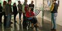A group of people listens attentively to a tour guide in a gallery. One person in a wheelchair observes alongside them. The scene is focused and engag