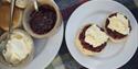 Two scones on a white plate topped with raspberry jam and clotted cream. Nearby, bowls of jam and cream are on a tartan blanket, evoking a cosy picnic