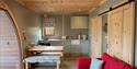 Cosy wood-paneled cabin interior with a compact kitchen, dining table, barn doors, and bright red sofa creating a warm, inviting feel.
