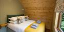 Cosy cabin bedroom with a neatly made bed, yellow knitted blanket, folded towels, patterned pillows, wood-paneled walls, and forest view.