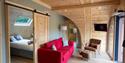 Cosy wood-paneled cabin interior with red sofa, armchair, loft bedroom, skylight, TV, and large windows; warm, inviting retreat.
