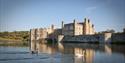 Medieval stone castle beside a calm moat, reflected in the water, with swans in front and a clear blue sky creating a tranquil scene.