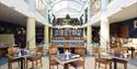 Bright, elegant restaurant interior with high, arched glass ceiling, wooden tables and chairs, neatly set for dining. Inviting and spacious ambiance.