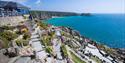 Coastal amphitheater with stone seats and colorful flowers overlooks a turquoise sea. Visitors enjoy the sunny day and picturesque ocean view.