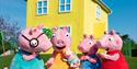 Peppa Pig characters in colorful outfits stand happily in front of a bright yellow house under a clear blue sky.