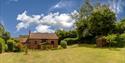 A charming brick cottage with a tiled roof sits amid lush greenery under a bright blue sky. A wooden gazebo stands to the right on a spacious lawn.