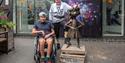 A young person in a wheelchair and an older person stand beside a bronze statue of Matilda. They are smiling, with a colorful, vibrant backdrop.