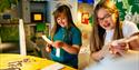 Two smiling girls engage in a crafting activity, cutting paper in a colorful, interactive exhibit space. The scene is lively and joyful.
