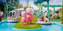 Colorful amusement park scene featuring large peppa pig statues on grassy island beside water ride with families in round boats. Playful, cheerful atm