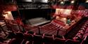Empty theater with red velvet seats and ornate balconies facing a dimly lit stage. Overhead lights cast a warm ambiance.