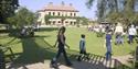 People walking along a path through a grassy field with a large house in the background