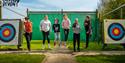 Six children joyfully jump in front of archery targets on a sunny day. The scene is lively and fun, conveying a sense of excitement and adventure.