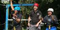 Three people wearing helmets and safety gear are cheering outdoors near a zipline platform. The scene is lively and joyful, with trees in the backgrou