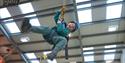 A child in green climbing gear and a blue helmet is happily suspended mid-air, gripping ropes in a bright indoor climbing facility.