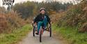 A person wearing a helmet joyfully rides an adaptive tricycle on a lush forest path, conveying a sense of freedom and adventure.