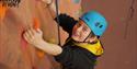 A person wearing a blue helmet and black sweater climbs an indoor rock wall with colorful holds, smiling confidently. The mood is adventurous and joyf