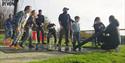A group of people outdoors, eagerly watching a science experiment involving a water rocket. The mood is joyful and anticipatory, with a sunny backdrop