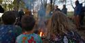 Two children in colorful pajamas sit by a campfire, surrounded by people in a wooded area. The scene feels warm and communal. Logo reads "Calvert Devo