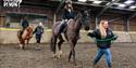 Two people ride horses in an indoor arena, led by instructors. The setting is calm and focused, highlighting equestrian training. "Calvert Devon" logo