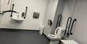changing places toilet with a toilet, handrails and cord. The floor is black and the wall is grey with white tiles.