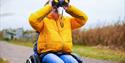 A child in a yellow coat and sat in a wheelchair with binoculars at WWT Arundel Wetland Centre