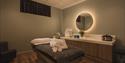 A serene spa room with a massage table topped by folded towels and slippers. Soft lighting, a glowing round mirror, and neatly arranged amenities crea