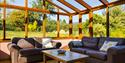 Cosy sunlit conservatory with brown leather sofas, decorative cushions, and a wooden coffee table. It overlooks a lush garden through large windows.